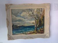 Framed landscape oil painting showing a lake, trees, and cloudy sky, ornate cream-colored frame