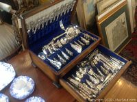 Wooden presentation case with hinged lid open, showing upper and lower compartments filled with flatware on blue velvet lining.
