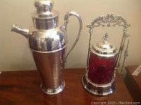 Side view of both items, showing the hammered silver cocktail shaker with lid and spout, and the red glass pickle jar with silver stand and tools.