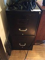 Front view of black metal filing cabinet with two drawers, metal handles, and label holders. Cabinet shows signs of wear.
