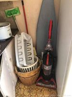Ironing board cover, laundry baskets, dish rack, and Dirt Devil upright vacuum placed together in a corner.