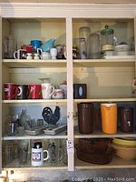 Wooden kitchen cabinet shelves filled with assorted kitchenware including mugs, glasses, pitchers, mixing bowls, and shakers.