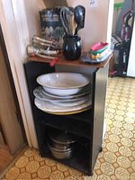 Photo of black wooden cart containing stacked plates, enamel bowl, metal cookware on bottom shelf, and black pitcher holding utensils on top shelf.