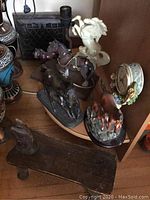 Photo of wood horse bench, horse lamp with bronze horse, white ceramic rearing horse statue, dark horse statues, and floral clock with horse figures on wooden shelf.