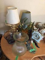 Two lamps with one having a shell base and cream-colored shade, blue lamp, eagle-themed clock, and several glass decor items including vases and bowls on a round wooden table.