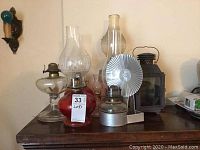 Seven oil lamps and one metal lantern arranged on a wooden surface, showing variety in glass shapes and colors, one lamp missing chimney