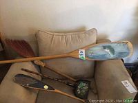 First photo showing the long wooden paddle with fish painting, bear paddle, small broom and decorative shovel placed on a beige armchair.