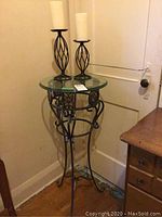 Glass top metal table accompanied by two metal candlesticks with pillar candles placed on top, displayed in corner near a white door.