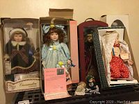 Photo showing five boxed porcelain dolls standing upright with visible certificates of authenticity on two dolls. Two mini porcelain dolls are in front sitting on the surface.