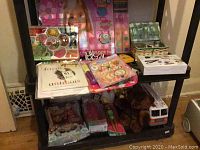 Overview of various toys and craft items on a black shelf including Barbie dolls, stuffed animals, craft kits, books, and Tonga fire engine toy.