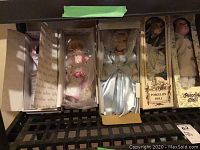 Five porcelain dolls in original boxes displayed side by side on a metal rack, some wrapping and protective packaging visible.