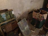 Photo showing gold metal-framed tray tables with wheels and framed paintings leaning against wall in basement area.