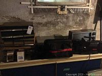 Photo showing a black CRT television, portable VHS player, black and red carrying bag, VHS tapes, and a printer on a shelf with dusty surroundings