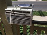 White KoolKing window air conditioner installed on wooden railing outdoors, showing front vents and control panel, with power cord hanging.