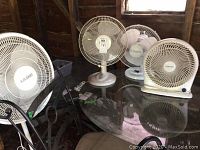 Four fans: three white table fans and one white floor fan, shown together on a table and chairs in a rustic setting.