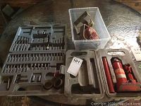 Photo showing the drill bit set case opened with various drill bits and accessories, a plastic container with metal jack stands and red reflector/light, and the Motomaster jack in its case.