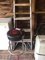 Photo showing Dodge logo cushioned black bar stool with chrome legs next to aluminum ladder against wooden wall.