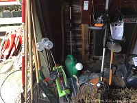 Photo showing Worx weed whacker and hedge trimmer with manual, multiple rakes, watering cans, and other garden tools arranged against a wall.