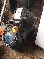 Full view of the air compressor showing the blue cylindrical tank, pressure gauges, yellow knob, and power cord.