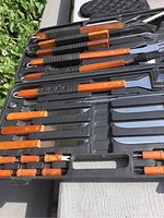 Overview of BBQ grilling tools and steak knives neatly arranged in a black carrying case, showing wooden handles and stainless steel parts.