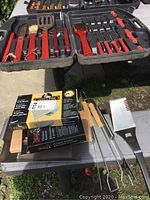 Full view of BBQ tools inside the plastic case showing red-handled spatula, fork, tongs, cooking brushes, skewers, carving knife and some boxed accessories below on table