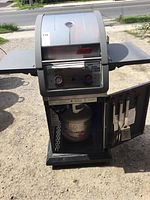 Full view of Coleman gas BBQ with side shelves and bottom cabinet open to show propane cylinder.