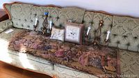 Overview of tapestry spread on green patterned sofa with two electric sconces and two embroidered pillows placed on top