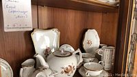 White Royal Copenhagen china set including teapot, square and oval plates, cups, saucers, creamer, and sugar bowl displayed on wooden cabinet shelves.