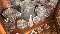 Photo shows various crystal and pressed glass serving and display items including covered jars, candy dishes, and small trays on wood furniture.