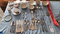 Overview of flatware, teacups, saucers, and serving utensils laid out on a blue cloth.