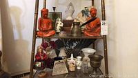 Wide shot of glass shelves holding the Asian inspired orange carved figurines, assorted porcelain and ceramic keepsakes, Asian fan, and the 1964 World's Fair tray