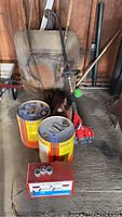 Photo showing rusty metal wheelbarrow, red weed wacker, and two empty metal gas cans on a floor inside a garage.