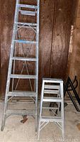 Photo showing two aluminum step ladders: one 6 foot with 8 steps and one 3 foot with 4 steps, standing on garage concrete floor against wood-paneled wall.