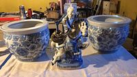 Two large blue and white fishbowl planters flanking the Asian fisherman figurine on a horse, all placed on a white cloth in a basement with yellow walls.