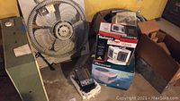 Wide view of boxed heater, humidifier, cords, and a large metal 20 inch window fan in basement area showing items in lot