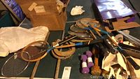 Photo of vintage wooden tennis racquets, hand weights, horseshoes, trophies, and game balls on a table.