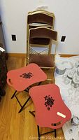 Five gold-tone metal folding chairs with brown velour seats and backrests stacked against the wall next to two red lacquered wood folding tray tables with floral accents on the floor in front.
