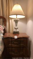 Wooden nightstand with two drawers and a glass table lamp on top, shown lit with fabric lampshade.