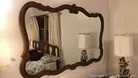 Full view of large rectangular mirror reflecting a bedroom with windows and bed visible. The framed mirror shows sculpted decorative detailing on wood.