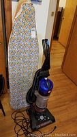 Hoover Wind Tunnel vacuum cleaner standing against an ironing board with a floral geometric patterned cover, showing wear and usage marks.