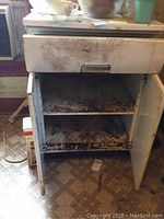 Front view of metal kitchen cabinet with single drawer and double doors open showing two rusted shelves inside.