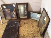 Four art pieces displayed on the floor against a wall including two framed prints, a framed mirror, and a wooden sign