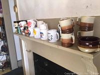 Row of over 25 assorted coffee mugs including printed logo mugs and two-tone brown and beige mugs with matching soup bowl