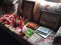 Photo of faux floral spray with large pink flowers, several framed prints (botanical and architectural themes), wooden plaques, and green glass ashtray arranged on a floral patterned couch