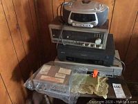Stack of electronics against wood paneled wall, including CD boom box, tuner, VCR, DVD player, and bundled cassette tapes.