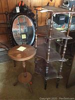 Wooden shelf unit with four curved shelves supported by spindles, a small round smoking table, and an oval mirror in the background.