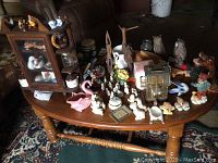 Overview of a wooden table with diverse collection of small figurines and decorative items placed on top, including animal figurines, planters, a clock, and a wooden cabinet with glass front.