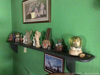 Side view of black wooden wall shelf with multiple figurines and a snow globe placed on top beneath a framed print on a green wall.