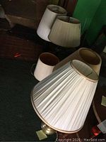 Six table lamps with various bases and an extra white pleated lampshade on dark carpeted floor.