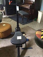 Photo of the stationary exercise bike showing the padded seat and part of the frame in a room with a piano in the background.